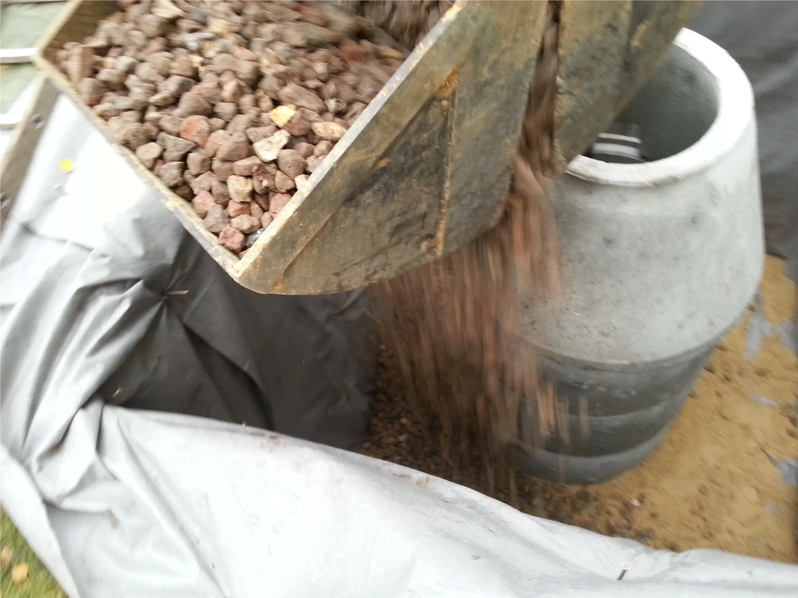 Drainagematerial wird neben dem Schachtkörper in die Baugrube eingebracht
